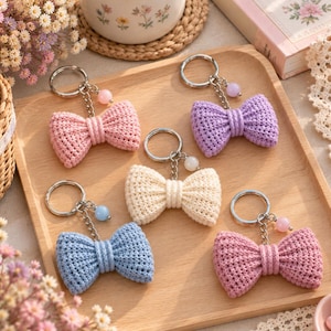 Pode incluir: Cinco porta-chaves de laço em croché em tons pastel, incluindo rosa claro, lavanda, creme e azul claro. Cada porta-chaves possui um anel de metal prateado, uma corrente curta e uma pequena conta redonda no centro do laço.
