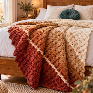 Könnte beinhalten: Eine gehäkelte Decke mit einem diagonalen Ombré-Muster, das von tiefem Rostrot zu gebranntem Orange, Hellbraun und Creme übergeht. Die Decke ist in einem strukturierten Granny-Square-Muster gefertigt und liegt über einer weißen Tagesdecke.