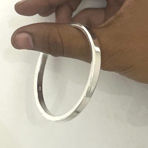 May include: A polished silver-tone bangle bracelet with a smooth, flat-edged band design. The metal surface is reflective and features a small, stamped hallmark on the inner side of the band, showcasing a clean, circular silhouette.