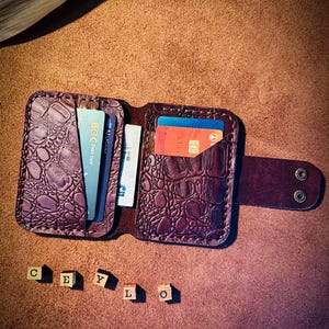 May include: A dark purple leather wallet with a textured crocodile skin pattern, featuring multiple card slots containing various debit and credit cards, a snap button closure strap, and five small wooden blocks spelling out CEYLO.