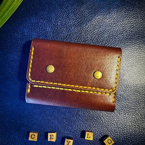 Può includere: Un portafoglio in pelle marrone con cuciture a contrasto gialle e due bottoni a pressione in metallo color ottone sulla patta anteriore. Sotto il portafoglio, cinque piccoli blocchi di legno mostrano le lettere C, E, Y, L, O.