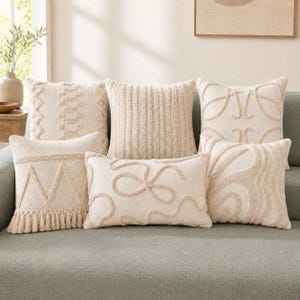 May include: A collection of six cream-colored throw pillows featuring various textured patterns including tufted zigzags, vertical stripes, swirling loops, and fringe details. The pillows are arranged in a group, showcasing different tactile designs.