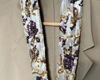 May include: A long, narrow scarf covered in silver, gold, and deep purple sequins arranged in a floral pattern. The scarf is draped over a light tan blazer hanging on a wooden hanger. The sequins create a reflective, textured surface.