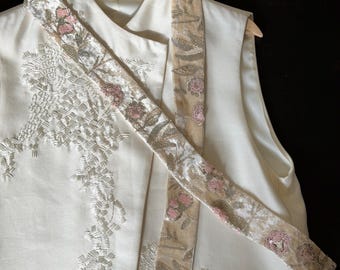 May include: A sleeveless off-white vest featuring intricate white thread embroidery and a long, attached sash. The sash is made of a light beige fabric adorned with floral patterns in pink thread, silver metallic accents, and small clear sequins.