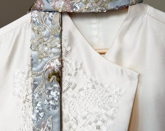 May include: An off-white blazer featuring a wrap-style front with intricate white beadwork embroidery. A detachable scarf in light blue mesh is draped over the collar, adorned with floral-patterned sequins and beads in silver, pink, and green.