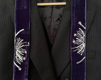 May include: A black pinstriped blazer layered with a long purple velvet scarf. The scarf features silver sequin embroidery in a firework-like pattern on both ends. The blazer has a notched lapel, a double-breasted front, and a black satin lining.