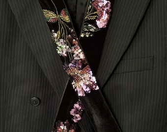 May include: A black pinstriped blazer featuring a double-breasted front with black buttons. Draped over the lapels is a long black velvet scarf adorned with intricate floral embroidery in shades of pink, purple, and green, accented with sequins.