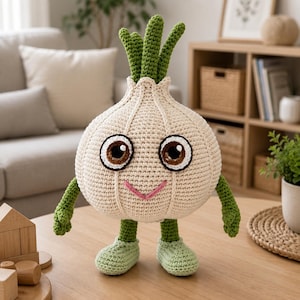 Puede incluir: Un peluche de ganchillo con forma de cabeza de ajo, cuerpo redondo color crema, tallos verdes como pelo, brazos verdes y pies verde claro. El ajo tiene grandes ojos marrones con reflejos blancos y una sencilla sonrisa bordada en color rosa.