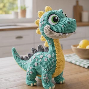Pode incluir: Um brinquedo de dinossauro em crochê com corpo verde-azulado, barriga amarela e espinhos dorsais amarelos. A criatura tem olhos grandes brancos e verdes, um sorriso largo com dentes brancos visíveis e manchas circulares cinza-claras no dorso.