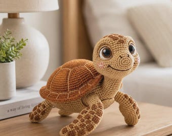 May include: A crocheted turtle plush toy featuring a light tan head and limbs with darker brown patches, a textured brown shell, large dark eyes, and small pink stitched cheeks. The turtle is depicted in a standing position with a smiling expression.