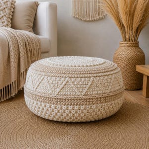 May include: A round, cream-colored crocheted pouf featuring a textured surface with a repeating geometric triangle pattern around the center. The pouf is constructed with thick yarn, showcasing a dense, knotted stitch design throughout its exterior.