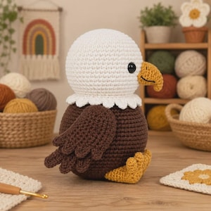 Puede incluir: Un juguete de peluche de águila calva hecho a ganchillo con cabeza blanca, cuerpo y alas de color marrón oscuro, pico amarillo y patas amarillas. El águila tiene ojos de plástico negro y está tejida con puntos visibles, sentada.