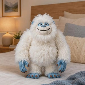 May include: A plush yeti toy featuring long white faux fur, a crocheted face with a wide smile, and light blue crocheted hands and feet with brown accents. The creature has small blue ears and a rounded, textured head with a soft, shaggy appearance.