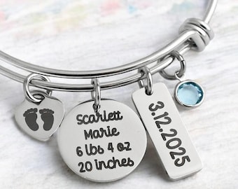 May include: Silver bangle bracelet with multiple charms. Charms include a heart with baby footprints, a round charm with a name and birth details, a rectangular charm with a date, and a small blue gemstone.