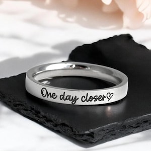 May include: A silver ring with the inscription "One day closer" and a small heart symbol.