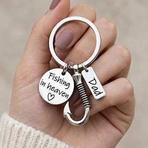 May include: A silver keychain with a fishing hook charm, a round charm with the text "Fishing in heaven" and a heart, and a rectangular charm with the word "Dad". The keychain is held in a hand.