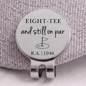 May include: A circular silver-toned metal golf ball marker with a clip, featuring the engraved text EIGHT-TEE and still on par, a small golf flag icon, and the text R.A. | 1946, attached to the brim of a textured light gray fabric cap.