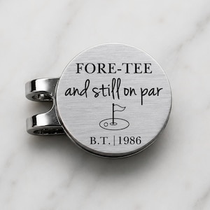 May include: A circular silver-toned metal golf ball marker with a clip attachment. The surface features the engraved text FORE-TEE and still on par, a small graphic of a golf flag in a hole, and the text B.T. | 1986 at the bottom.