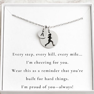May include: A silver necklace with two charms. One charm is a small circle with the letter "A", and the other is a larger circle with a silhouette of a person running. The necklace is presented in a white box with text.