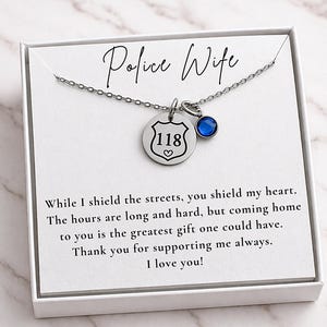 K&ouml;nnte beinhalten: Eine silberne Halskette mit einem Anh&auml;nger "Police Wife". Der Anh&auml;nger hat ein Abzeichen mit der Nummer "118" und einen blauen Edelstein. Die Karte lautet "While I shield the streets, you shield my heart."