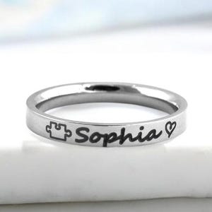 May include: A silver ring engraved with the name "Sophia", a puzzle piece and a heart symbol.
