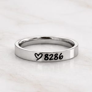 Puede incluir: Un anillo plateado con el número "8286" y un símbolo de corazón grabado en negro. El anillo es circular y descansa sobre una superficie de mármol blanco.