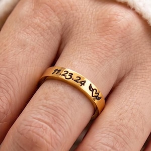 Peut inclure: Une bague dorée gravée de la date "11.23.24" et d'un motif cœur et feuille. La bague est portée au doigt, sur un fond de pull blanc et de fleurs floues. La bague est une simple alliance.
