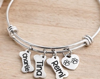 May include: Silver-toned charm bracelet with adjustable band. Features bone-shaped charms engraved with the names "Charlie", "Tina", and "Remi", plus a heart charm with paw prints. The bracelet is displayed on a wooden surface.