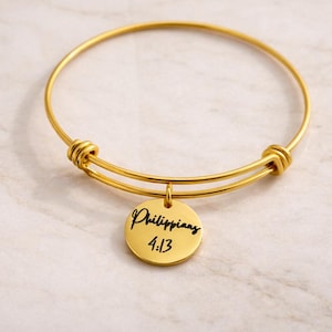 May include: A gold-colored bangle bracelet with a round charm. The charm is engraved with the word "Philippians" and the numbers "4:13" in a handwritten style font. The bracelet is on a marble surface.