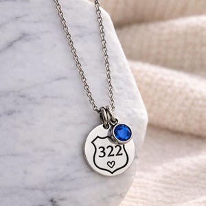K&ouml;nnte beinhalten: Silberfarbene Halskette mit einem runden Anh&auml;nger mit der Nummer "322" in einem Schild und einem kleinen Herz darunter. Ein blauer Edelstein ist &uuml;ber dem Anh&auml;nger angebracht. Die Halskette wird auf einer wei&szlig;en Oberfl&auml;che pr&auml;sentiert.