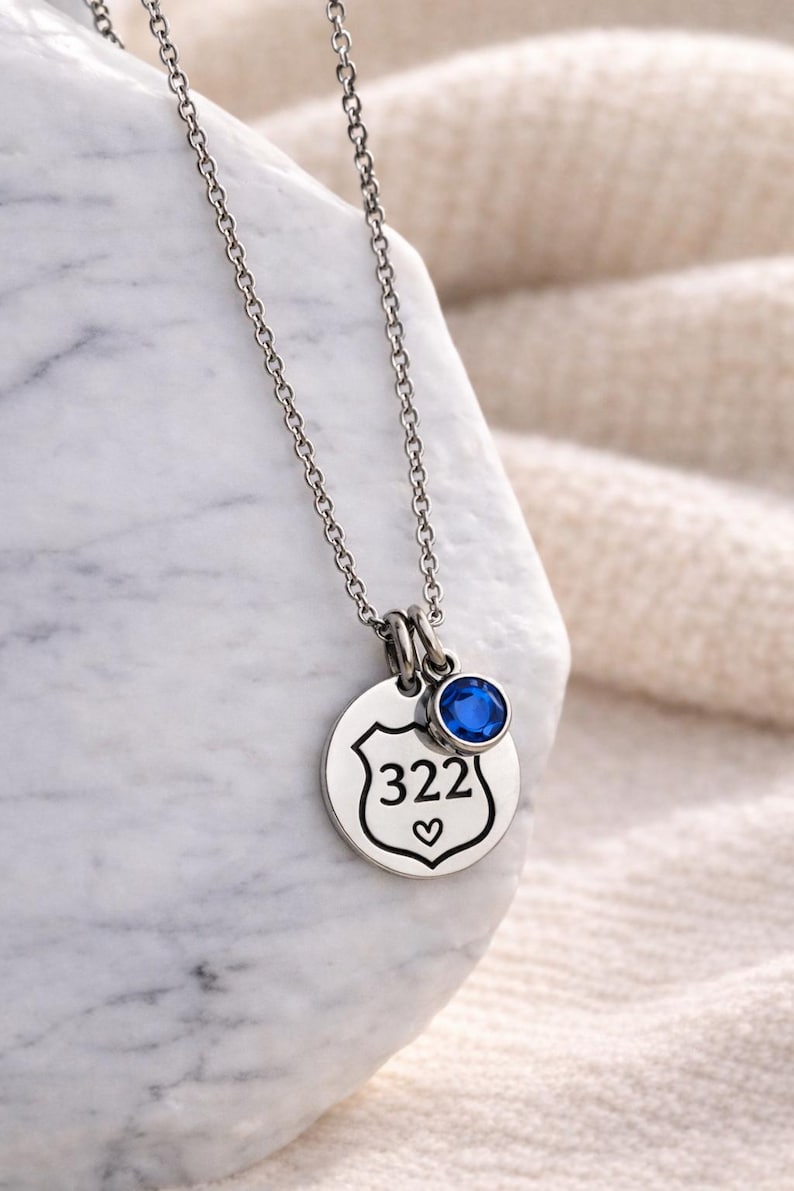 K&ouml;nnte beinhalten: Silberfarbene Halskette mit einem runden Anh&auml;nger mit der Nummer "322" in einem Schild und einem kleinen Herz darunter. Ein blauer Edelstein ist &uuml;ber dem Anh&auml;nger angebracht. Die Halskette wird auf einer wei&szlig;en Oberfl&auml;che pr&auml;sentiert.