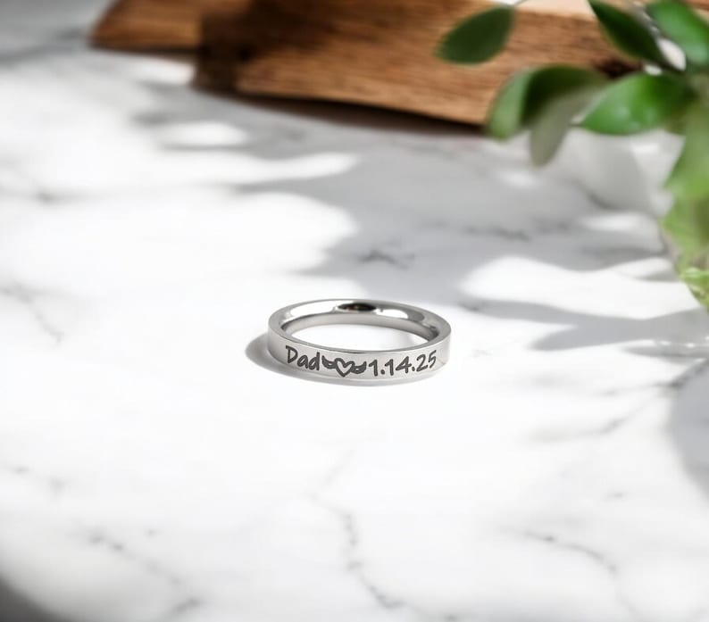May include: A silver ring with the inscription "Dad" followed by a heart symbol and the date "1.14.25".