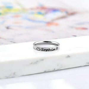 Pode incluir: Um anel de prata com o nome "Sophia" gravado, juntamente com um pequeno cora&ccedil;&atilde;o e um s&iacute;mbolo de pe&ccedil;a de quebra-cabe&ccedil;a.