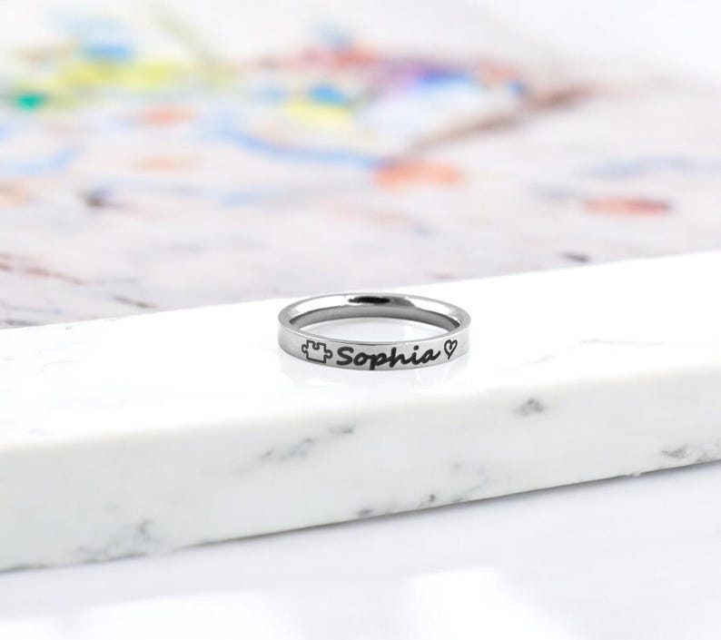 Pode incluir: Um anel de prata com o nome "Sophia" gravado, juntamente com um pequeno cora&ccedil;&atilde;o e um s&iacute;mbolo de pe&ccedil;a de quebra-cabe&ccedil;a.