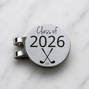 Peut inclure: Marqueur de balle de golf argenté avec clip. Le marqueur porte l'inscription "Class of 2026" en lettres cursives et bâtons noirs, avec des clubs de golf croisés en dessous. Les initiales "J.S." sont en bas.