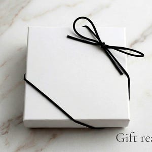 K&ouml;nnte beinhalten: Eine quadratische, wei&szlig;e Geschenkbox, die mit einer schwarzen Kordelschleife gebunden ist. Die Box befindet sich auf einer Marmoroberfl&auml;che. Der Text "Gift ready" ist unten rechts in Schwarz gedruckt.