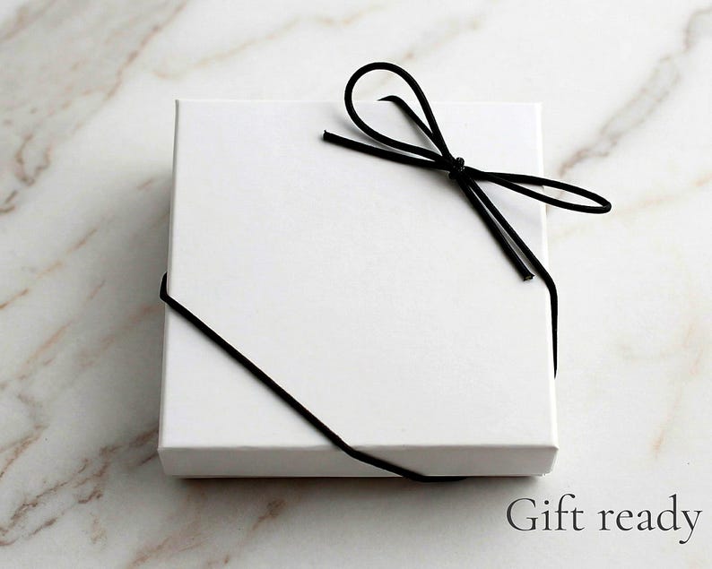 K&ouml;nnte beinhalten: Eine quadratische, wei&szlig;e Geschenkbox, die mit einer schwarzen Kordelschleife gebunden ist. Die Box befindet sich auf einer Marmoroberfl&auml;che. Der Text "Gift ready" ist unten rechts in Schwarz gedruckt.