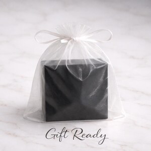 Puede incluir: Una caja de regalo negra cuadrada est&aacute; dentro de una bolsa de organza blanca transparente, atada con una cinta a juego. Las palabras "Gift Ready" est&aacute;n escritas en una elegante escritura en la parte inferior de la imagen. El fondo es una superficie similar al m&aacute;rmol.