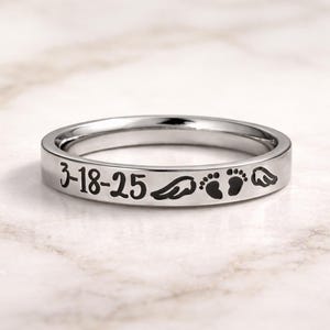 Może przedstawiać: Srebrny pierścionek z wygrawerowanym tekstem i symbolami. Tekst brzmi "3-18-25", a następnie skrzydło, dwa odciski st&oacute;p dziecka i kolejne skrzydło. Pierścionek jest prezentowany na marmurowej powierzchni.