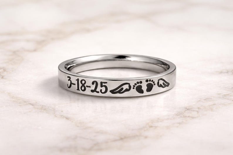 Może przedstawiać: Srebrny pierścionek z wygrawerowanym tekstem i symbolami. Tekst brzmi "3-18-25", a następnie skrzydło, dwa odciski st&oacute;p dziecka i kolejne skrzydło. Pierścionek jest prezentowany na marmurowej powierzchni.