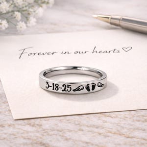 Może przedstawiać: Srebrny pierścionek z wygrawerowaną datą "3-18-25", skrzydłami anioła i odciskami st&oacute;p dziecka. Pierścionek leży na kartce z odręcznym napisem "Forever in our hearts". W tle znajduje się długopis.