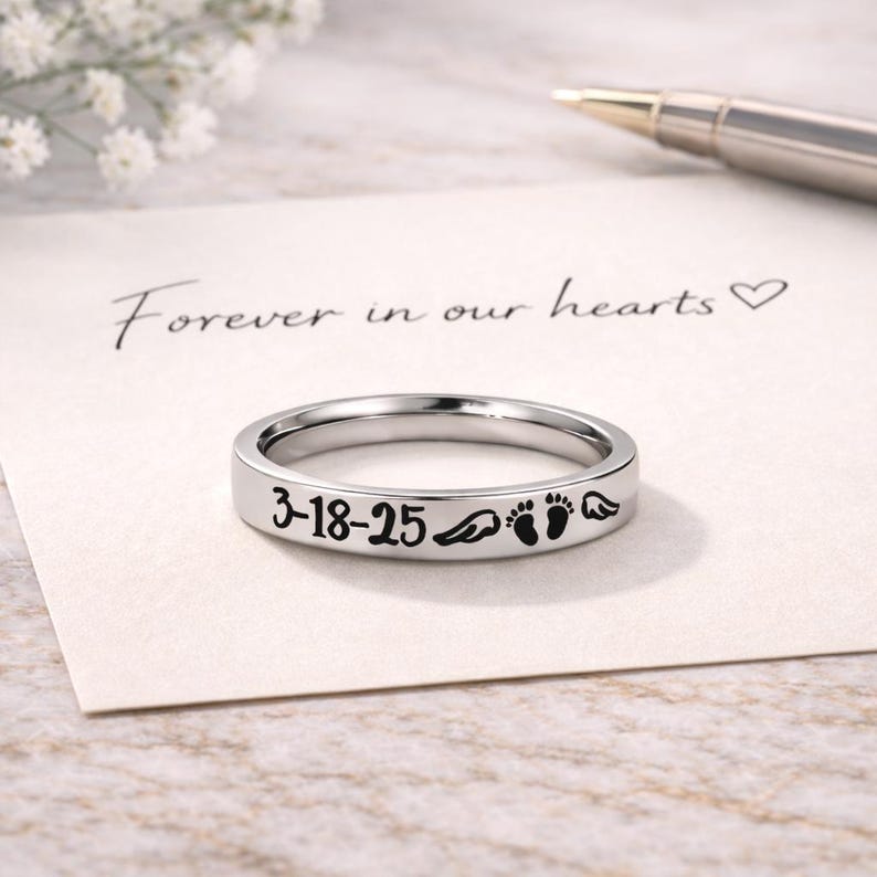 Może przedstawiać: Srebrny pierścionek z wygrawerowaną datą "3-18-25", skrzydłami anioła i odciskami st&oacute;p dziecka. Pierścionek leży na kartce z odręcznym napisem "Forever in our hearts". W tle znajduje się długopis.
