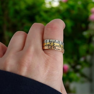 May include: Three stacked silver rings with engraved messages: "be still", "just breathe", and "let them" with a heart symbol after each message.