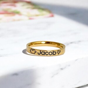 Pode incluir: Um anel dourado com o nome "Jacob" gravado, juntamente com uma pe&ccedil;a de quebra-cabe&ccedil;a e um s&iacute;mbolo de cora&ccedil;&atilde;o.