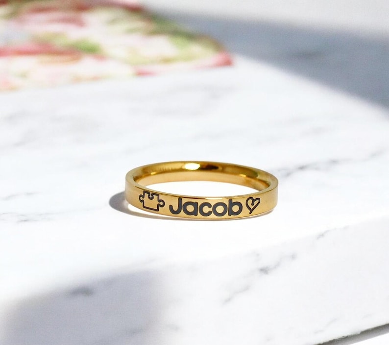 Pode incluir: Um anel dourado com o nome "Jacob" gravado, juntamente com uma pe&ccedil;a de quebra-cabe&ccedil;a e um s&iacute;mbolo de cora&ccedil;&atilde;o.