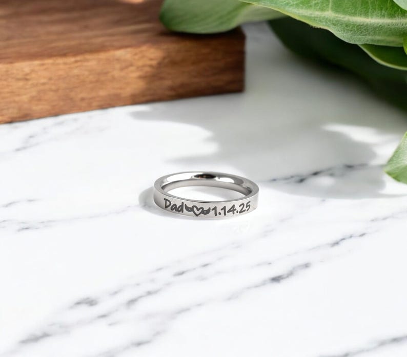 May include: A silver ring with the inscription "Dad" followed by a heart symbol and the date "1.14.25".