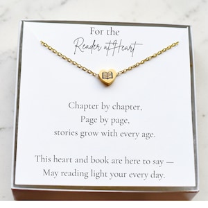 Pode incluir: Um colar dourado com um pingente em forma de coração com um ícone de livro. O colar é exibido em um cartão branco com o texto "For the Reader at Heart" e um poema sobre leitura.