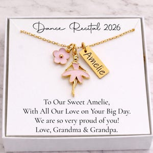 May include: A gold-colored necklace with a ballerina charm, a pink flower charm, and a rectangular tag with the name "Amelie". The necklace is presented in a white box with the text "Dance Recital 2026".
