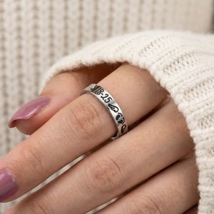 Może przedstawiać: Srebrny pierścionek z wygrawerowanym napisem "3-18-25" i małymi odciskami st&oacute;p. Pierścionek jest noszony na palcu. Pierścionek to biżuteria.