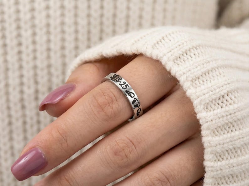 Może przedstawiać: Srebrny pierścionek z wygrawerowanym napisem "3-18-25" i małymi odciskami st&oacute;p. Pierścionek jest noszony na palcu. Pierścionek to biżuteria.